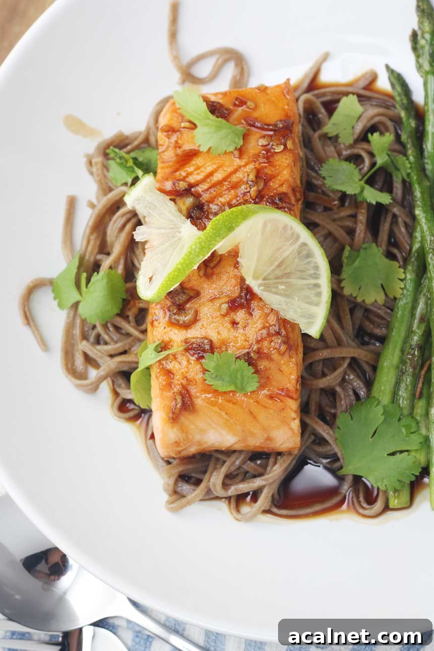 An overhead view of a white dish of maple teriyaki salmon with a twisted slice of lime on top and with soba noodles and asparagus.