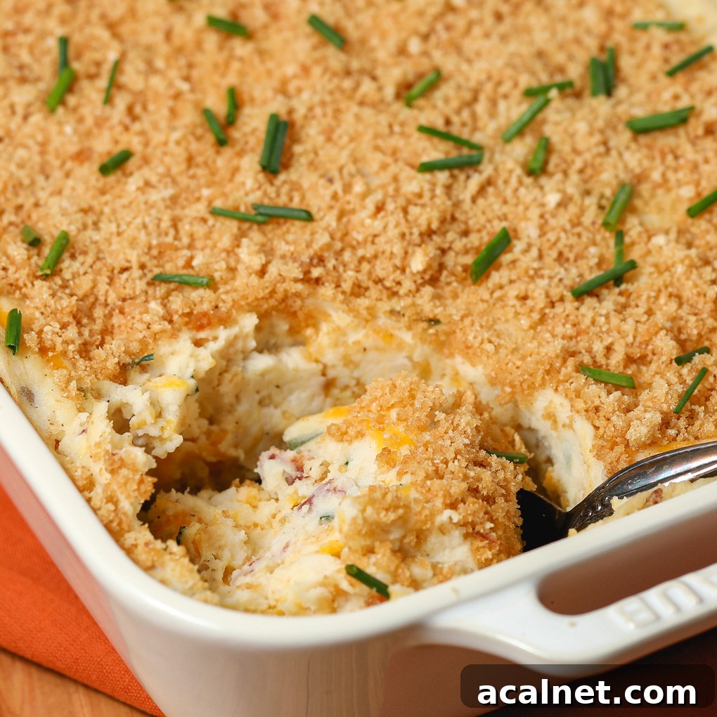 A Gluten and Dairy Free Twice Baked Potato Casserole with a golden breadcrumb topping, garnished with chopped chives. A portion has been scooped out, revealing a creamy interior with bits of bacon and cheese.