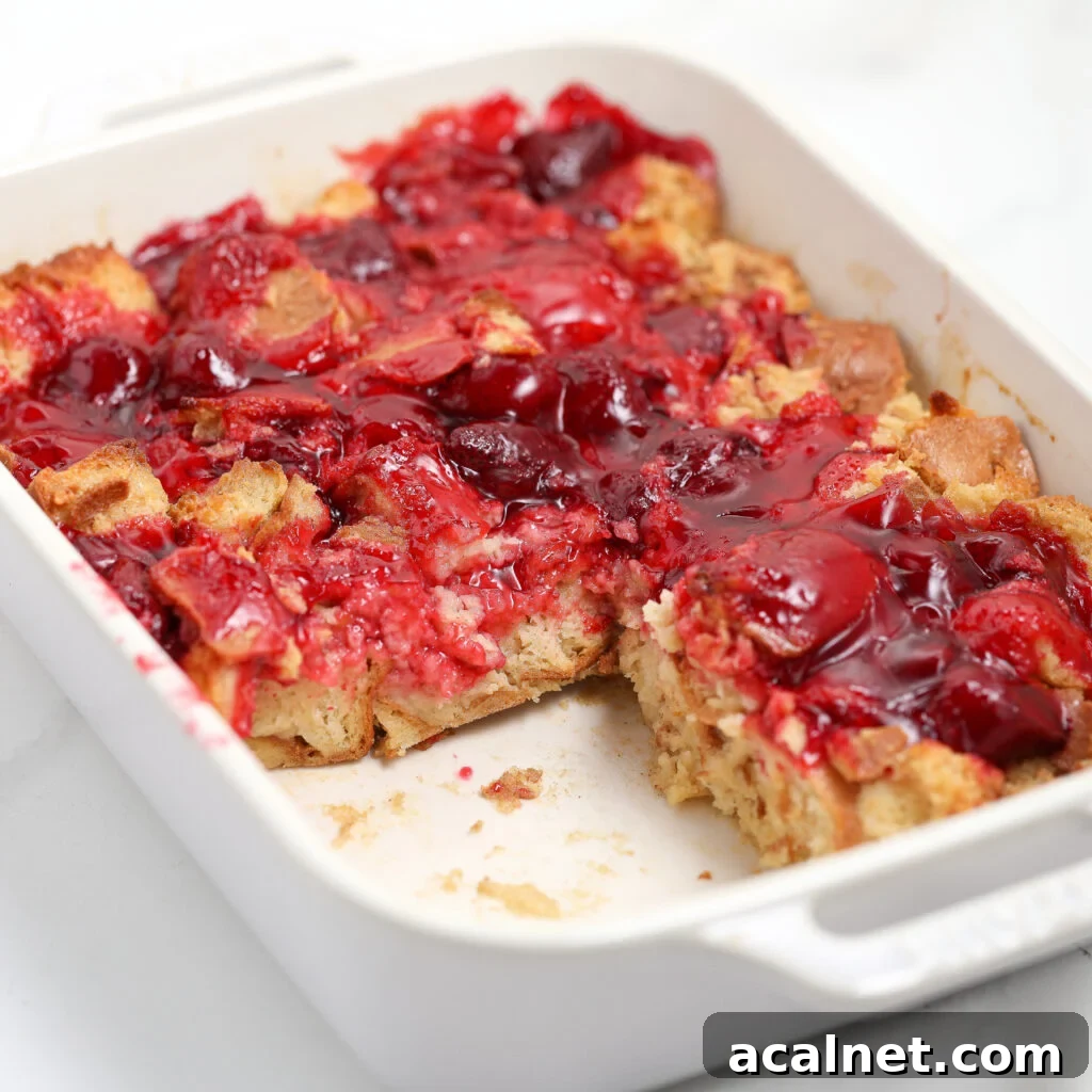 A white baking dish filled with cherry bread pudding; some pieces have been served, revealing chunks of bread topped with bright red cherry filling, similar to a strawberry french toast casserole in appearance and appeal.