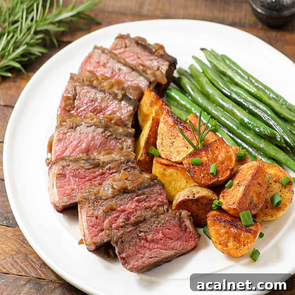 A white plate with sliced medium-rare steak topped with onions, roasted potato wedges garnished with chives, and green beans—perfect for a Father's Day feast. A sprig of rosemary rests on the table nearby.