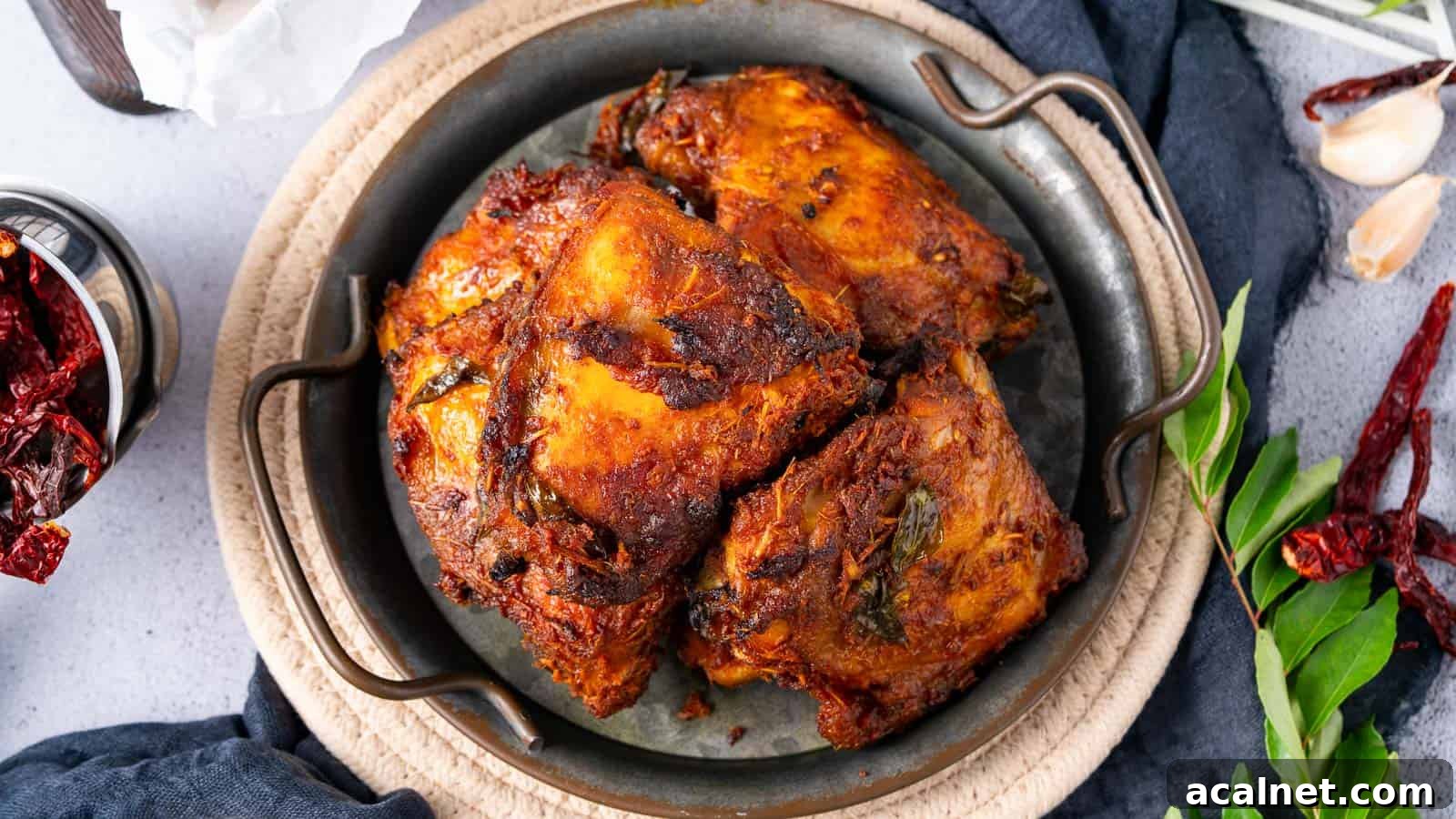 29 Chicken Dinners That Keep Coming Back to Our Weekly Menu 3 A plate of spicy, roasted chicken pieces garnished with herbs, surrounded by dried red chilies, garlic cloves, and green leaves on a light surface.
