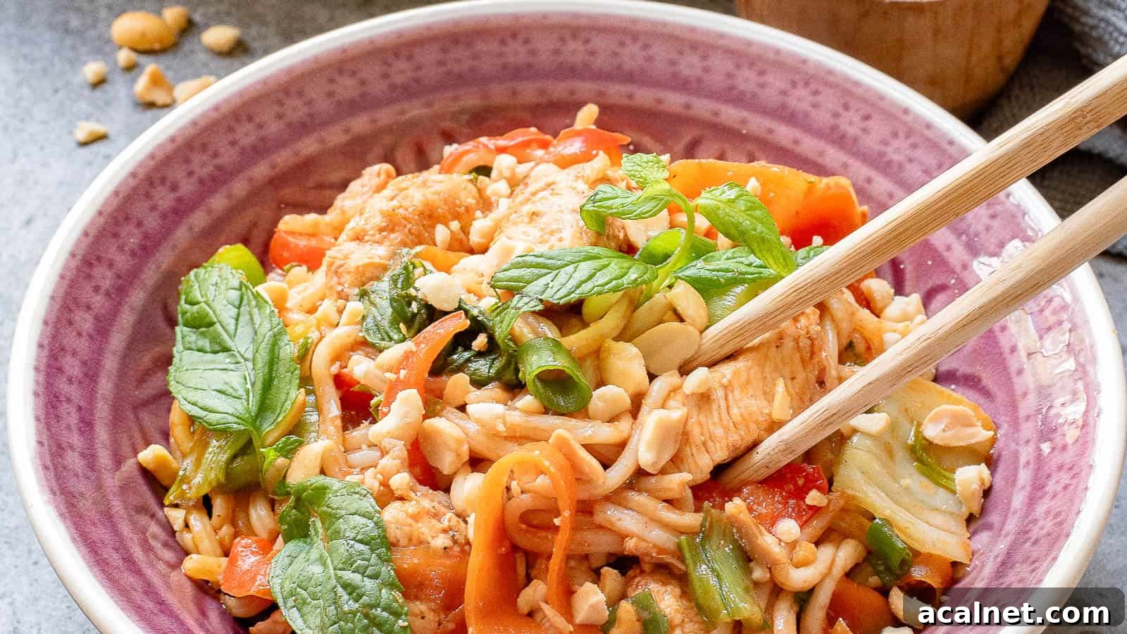 29 Chicken Dinners That Keep Coming Back to Our Weekly Menu 30 A bowl of noodle stir-fry topped with vegetables, chicken, crushed peanuts, and fresh mint leaves. Chopsticks rest on the side of the bowl, which is placed on a textured surface with a small container in the background.