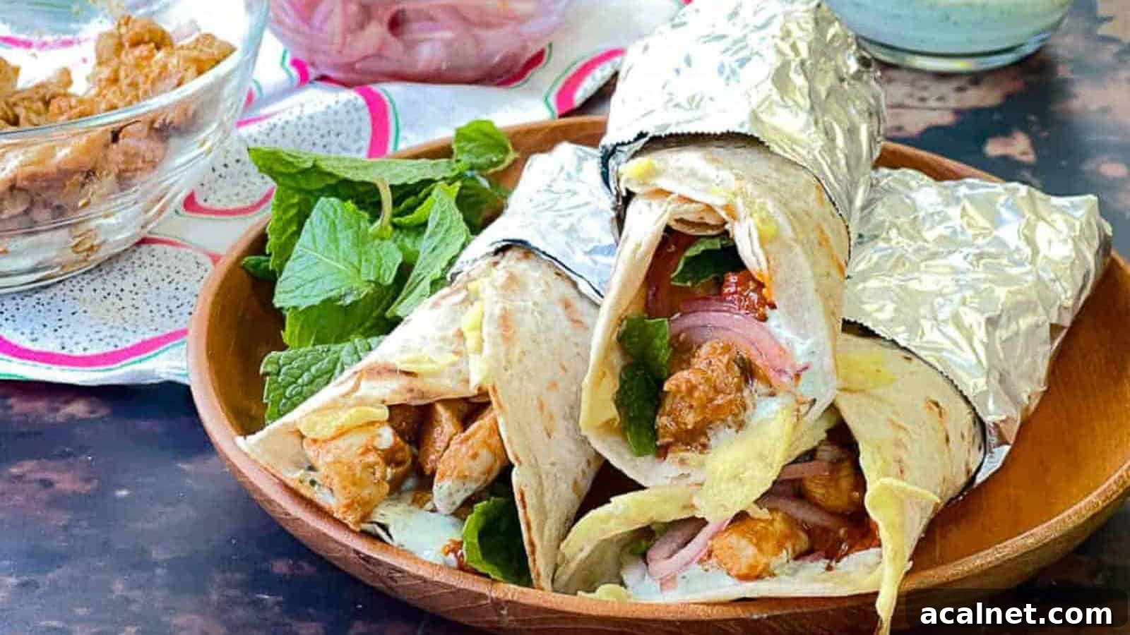 29 Chicken Dinners That Keep Coming Back to Our Weekly Menu 4 Low angle shot of 3 chicken kathi rolls wrappedin foil on a wooden plate with fresh mint leaves.