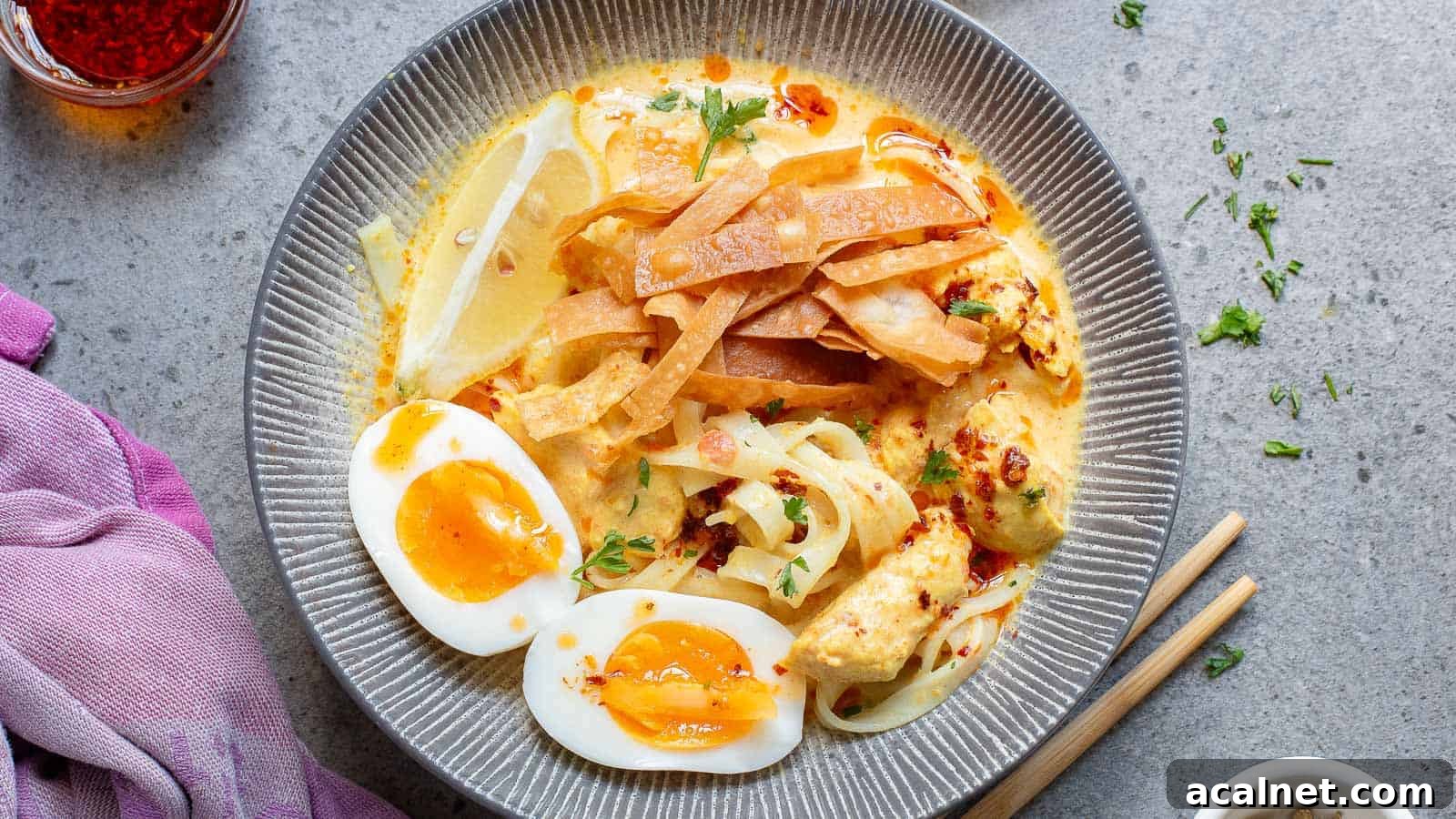 29 Chicken Dinners That Keep Coming Back to Our Weekly Menu 9 A gray bowl filled with a creamy noodle soup topped with sliced boiled eggs, crispy fried noodles, lemon wedges, and herbs. A napkin, chopsticks, and a small jar of spices are nearby on the gray surface.