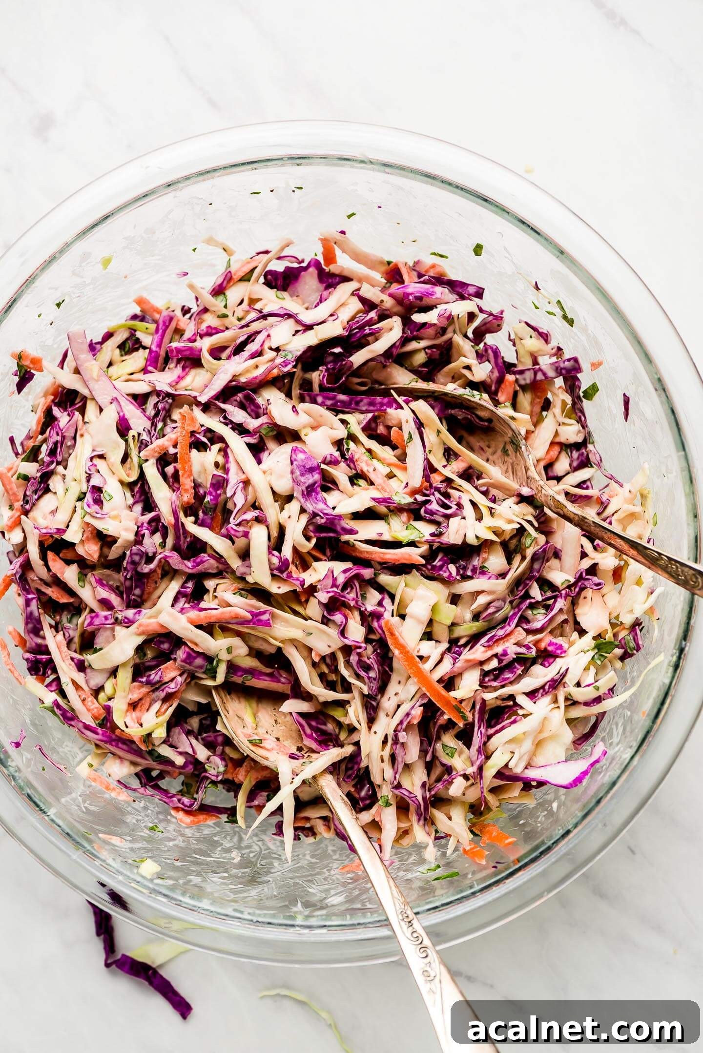A glass mixing bowl of Coleslaw with two serving spoons.
