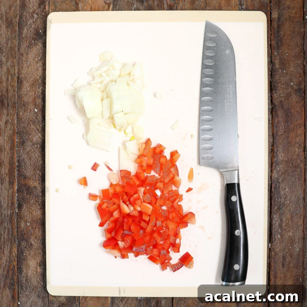 A chopping board on a wooden surface holds chopped onions, diced red bell peppers, and a large kitchen knife with a black handle—ingredients ready for making Pork Meatloaf with Gravy.