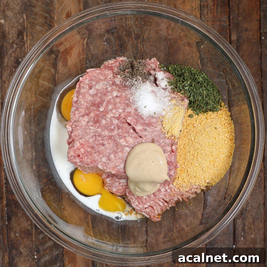 A glass bowl on a wooden surface holds ingredients for Pork Meatloaf with Gravy: ground meat, eggs, milk, mustard, breadcrumbs, dried herbs, salt, and pepper—all ready to be mixed.
