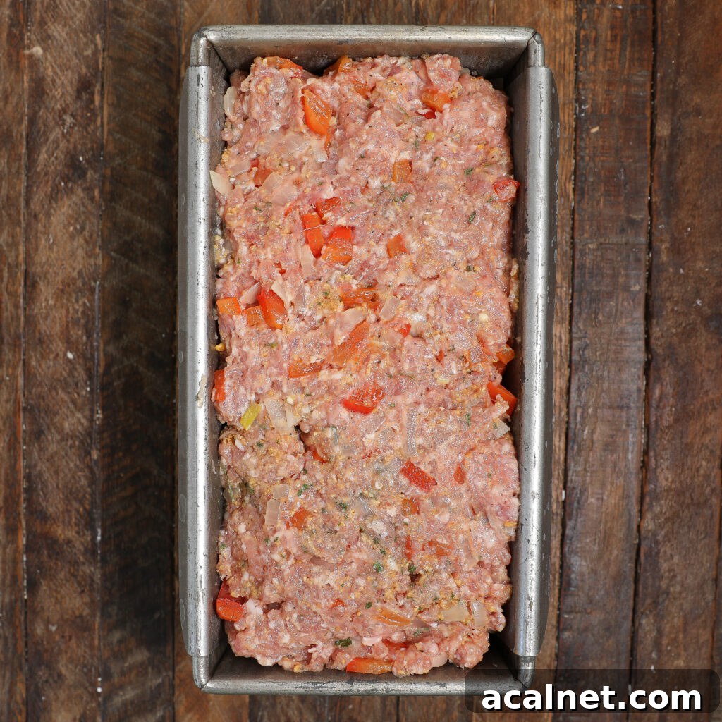 A metal loaf pan filled with uncooked Pork Meatloaf with Gravy mixture, including ground meat, chopped red bell peppers, and onions, sits on a rustic wooden surface.