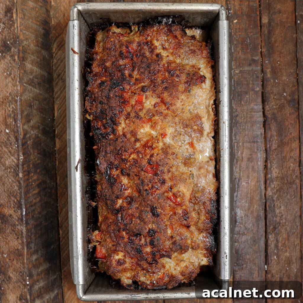 A browned, cooked Pork Meatloaf with Gravy and bits of vegetables is shown in a rectangular metal loaf pan, resting on a rustic wooden surface.