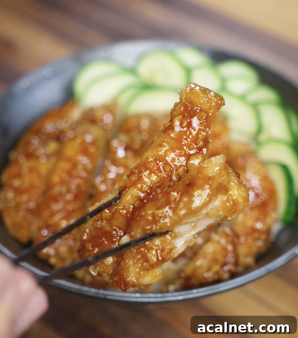 A piece of crispy chicken coated in honey garlic glaze is held up with chopsticks, highlighting the juicy interior and glossy, crunchy exterior.
