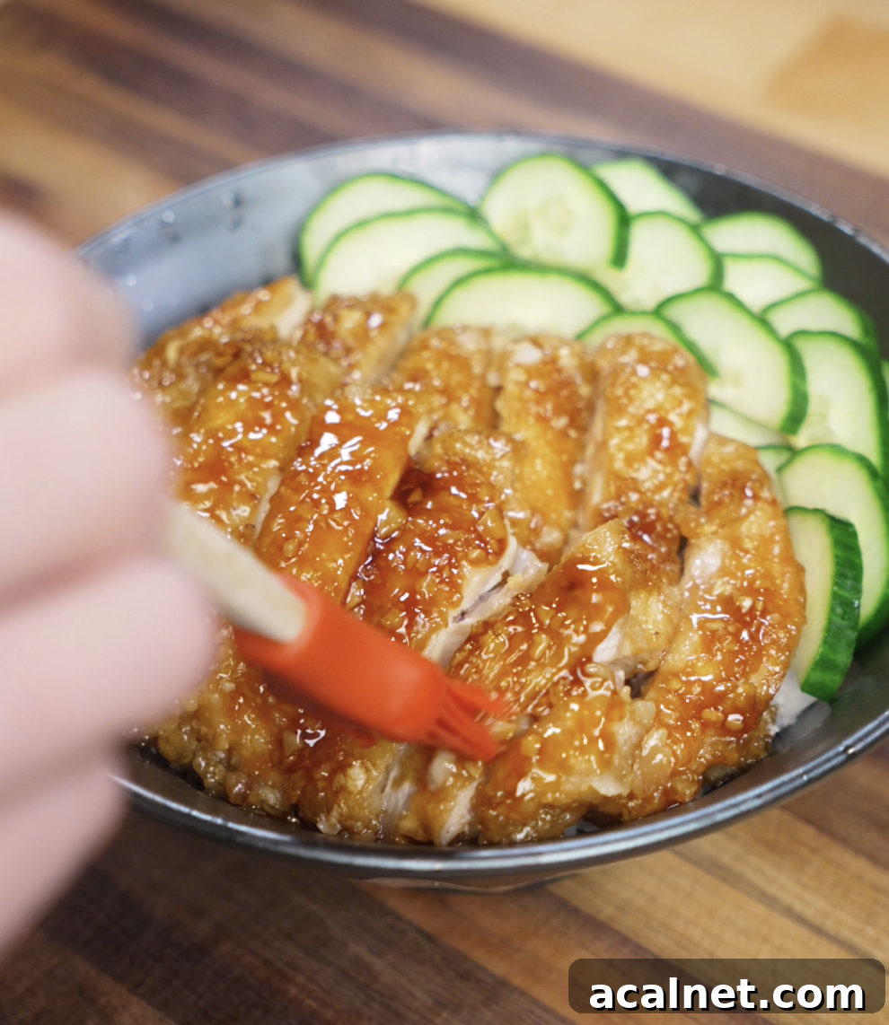 A hand uses a red brush to apply extra honey garlic glaze over sliced crispy chicken thighs, adding shine and flavor to the final rice bowl topped with cucumbers.