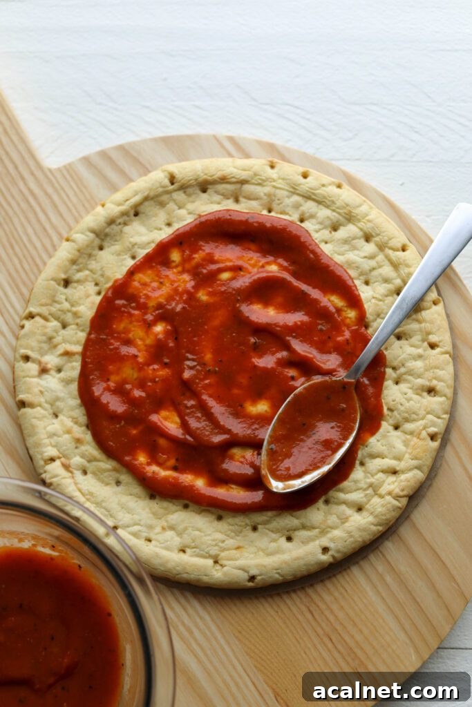 Banza chickpea crust with pizza sauce and a spoon on wooden board by Flora & Vino