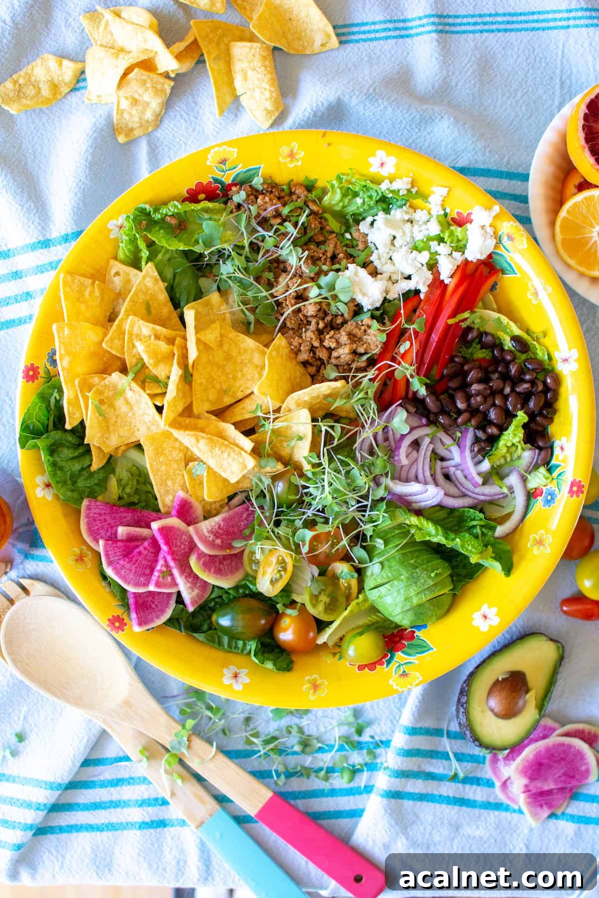 A yellow bowl filled with taco salad sitting on a blue tablecloth with a pink-handled spoon next to it and several chips. 