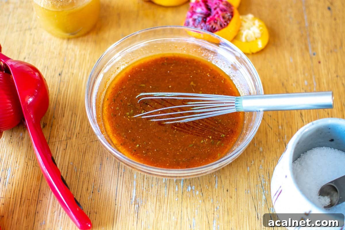 A glass bowl sitting on a wood table with the dressing for taco salad in it and a wire whisk in the bowl with citrus around. 