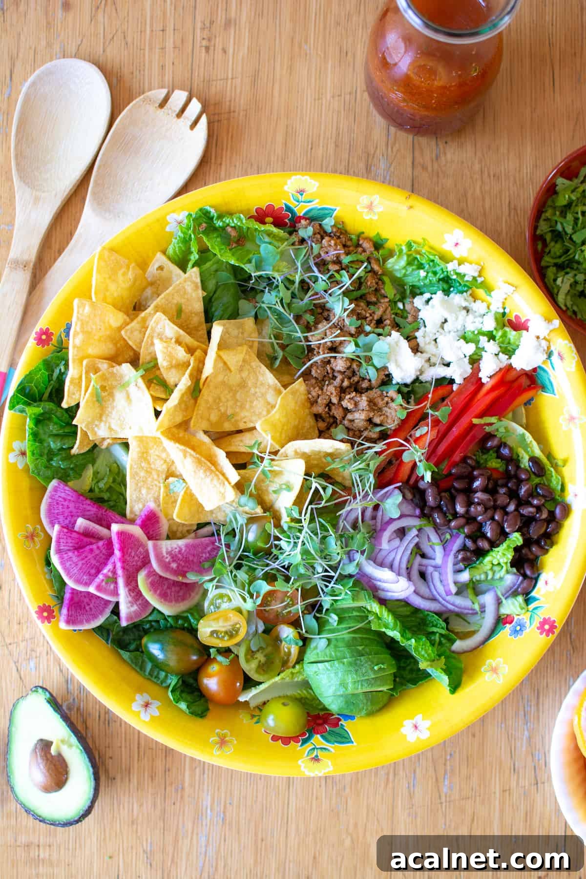 A big yellow bowl filled with taco salad ingredients on a wood table with a carafe of salad dressing next to it. 
