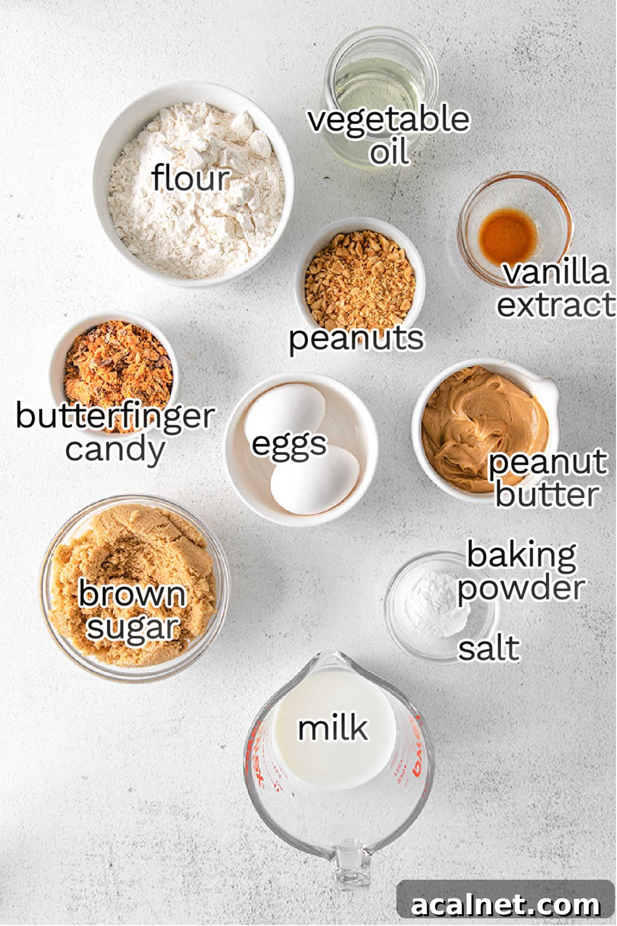 A flat lay of ingredients for baking peanut butter cupcakes, including flour, vegetable oil, vanilla extract, peanuts, peanut butter, eggs, Butterfinger candy, brown sugar, baking powder, salt, and milk.