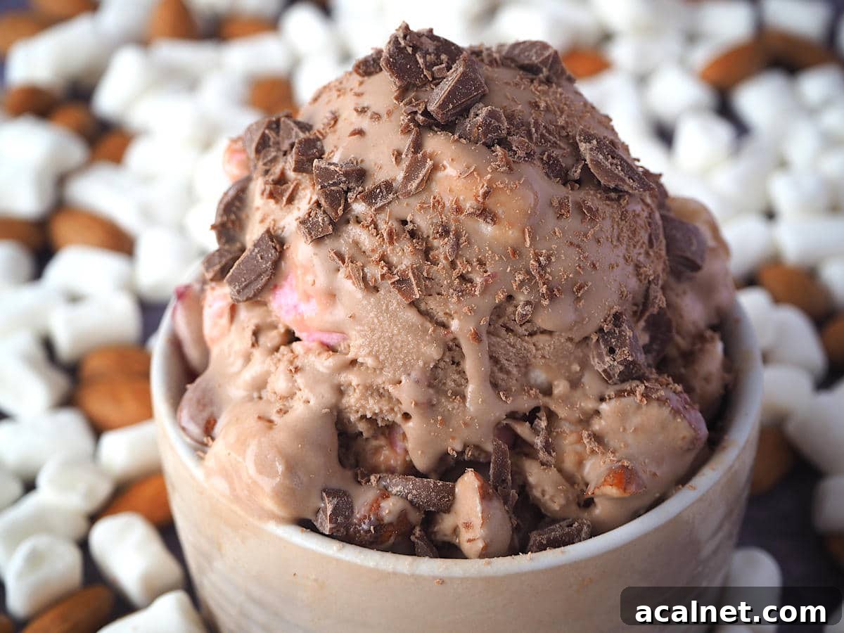 Close up scoop of rocky road ice cream with chopped chocolate sprinkled on top and ice cream drip, in a brown bowl, with marshmallows and almonds in the background.