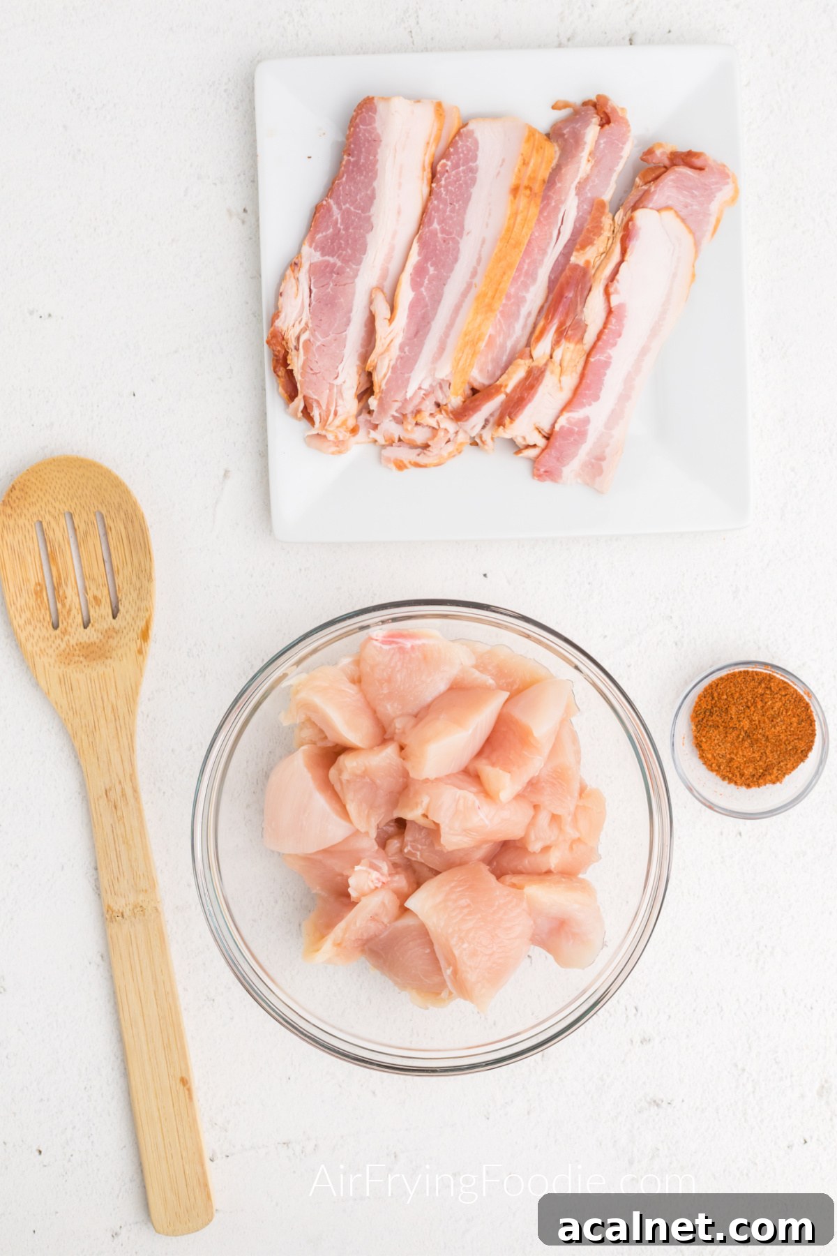 Bacon on a white plate, seasonings in a small container, and chicken pieces in a bowl. 