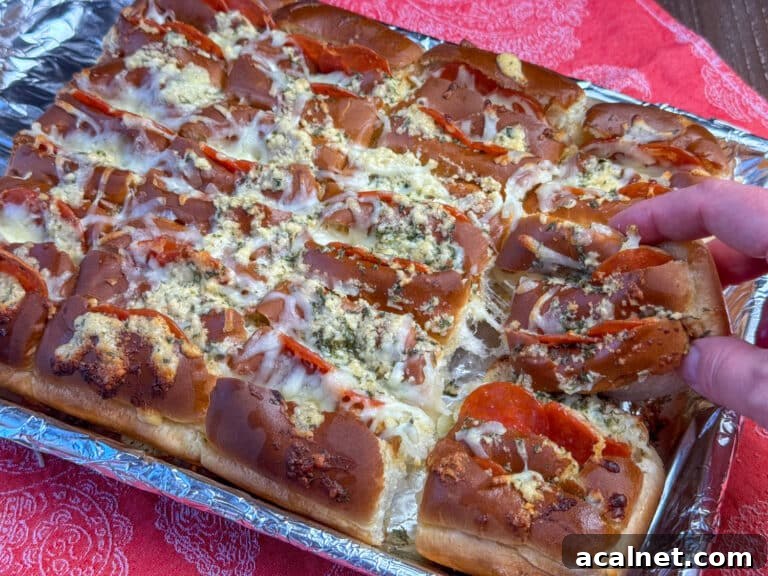 Hand pulling a Hawaiian roll stuffed with pepperoni and cheese off of a baking sheet full of rolls.