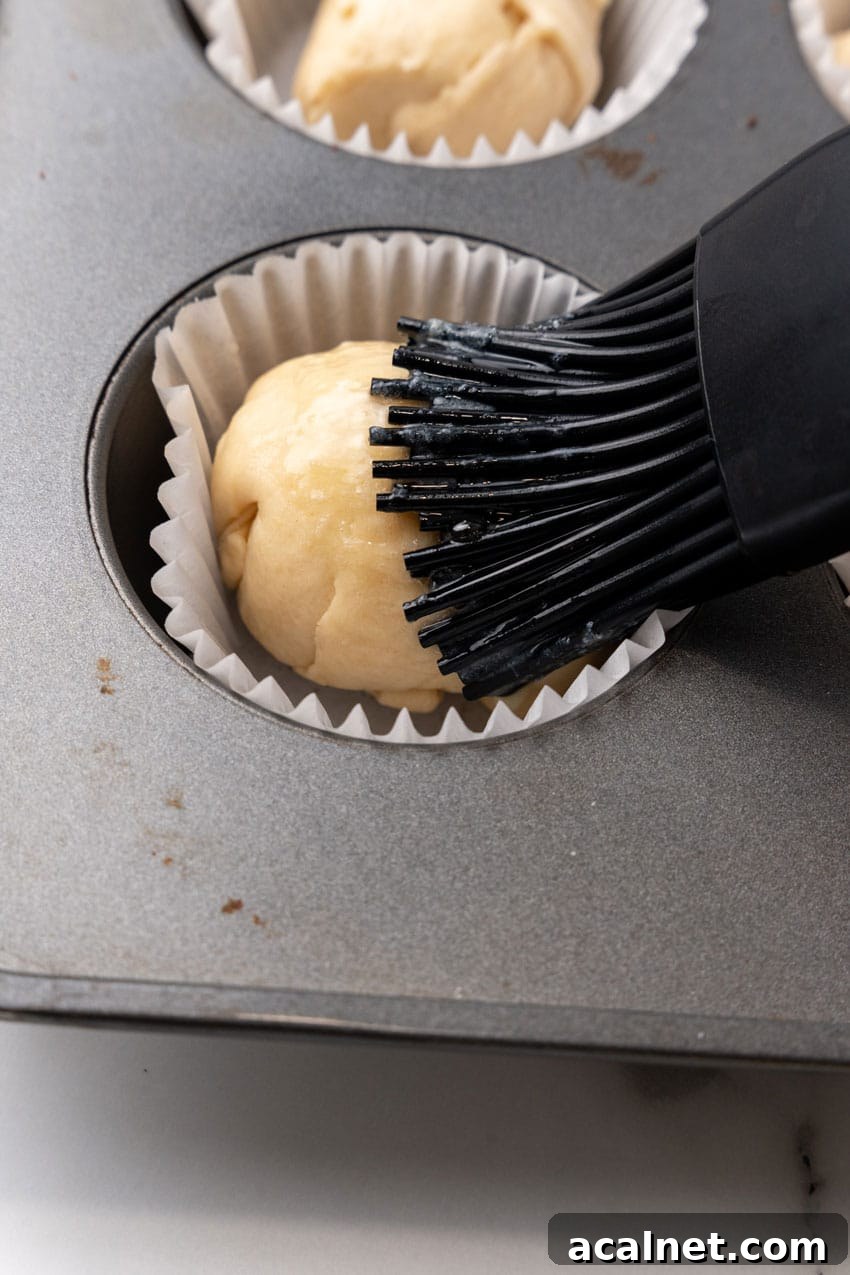 a pastry brush rubbing melted butter over an unbaked resurrection roll in a metal muffin pan