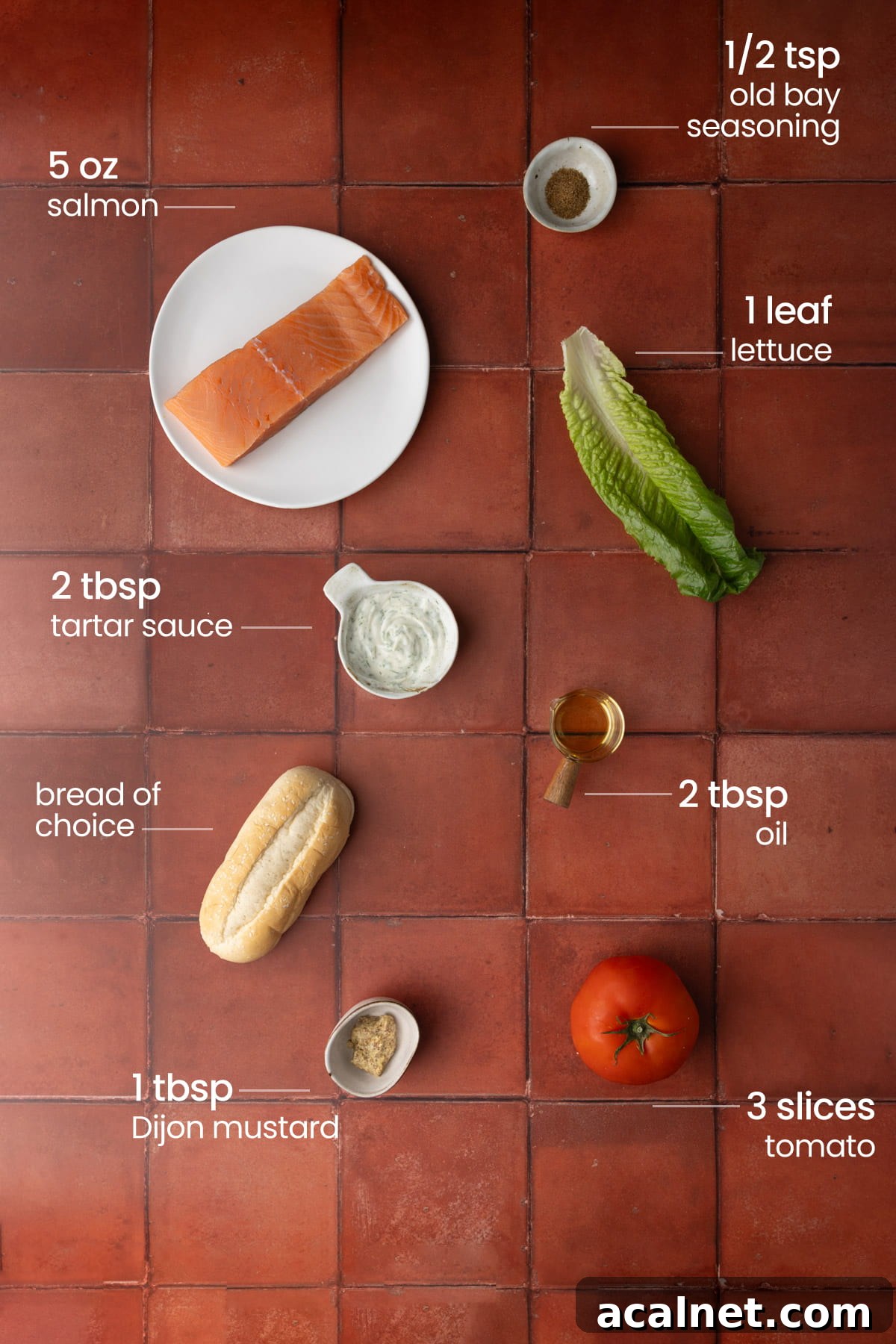 Overhead view of all the ingredients needed for a pan-fried salmon sandwich - old bay seasoning, lettuce, salmon, tartar sauce, oil, bread, Dijon mustard, and tomato.