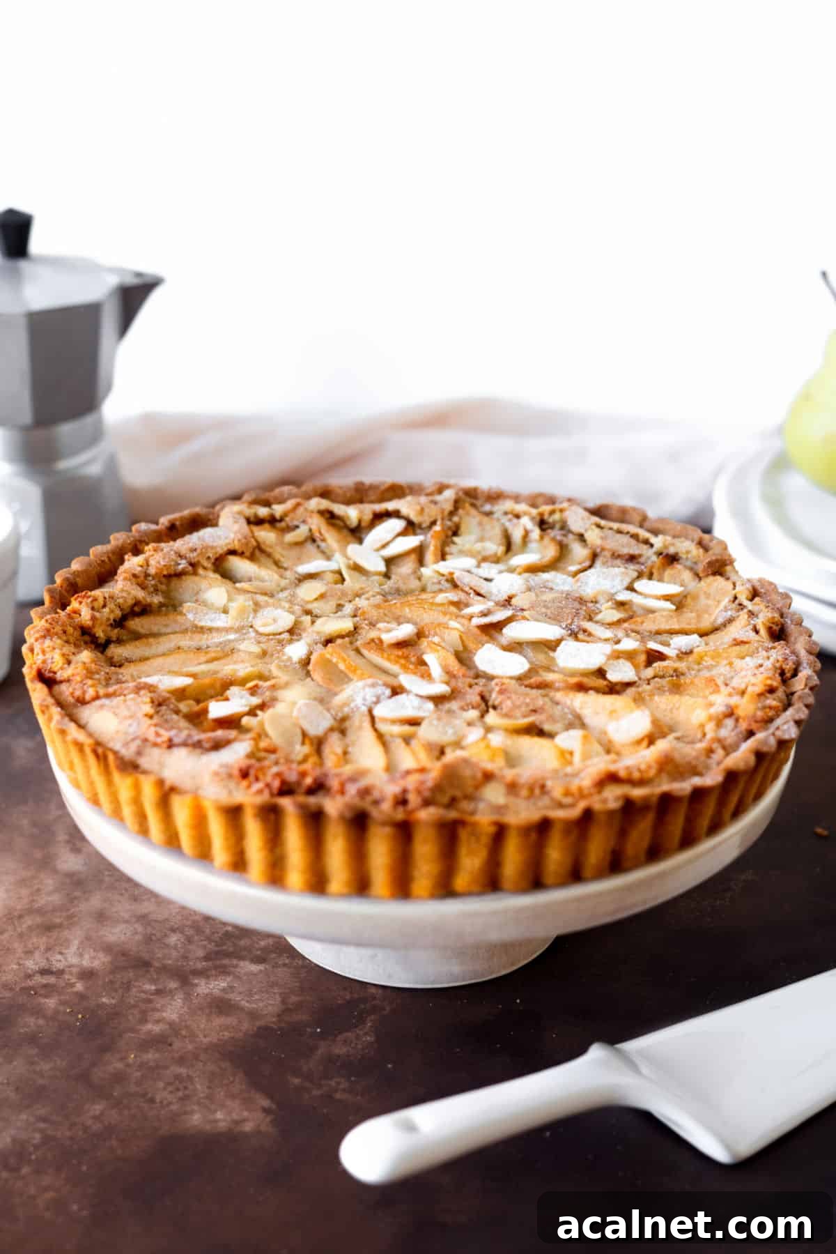 Round pear almond tart on a white cake stand. Brown surface, white background.