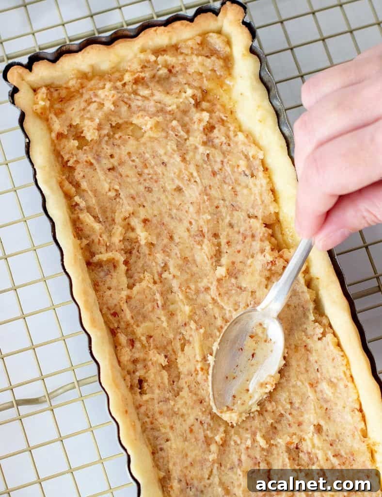 Hand spreading almond cream on rectangular pie crust on a wire rack.