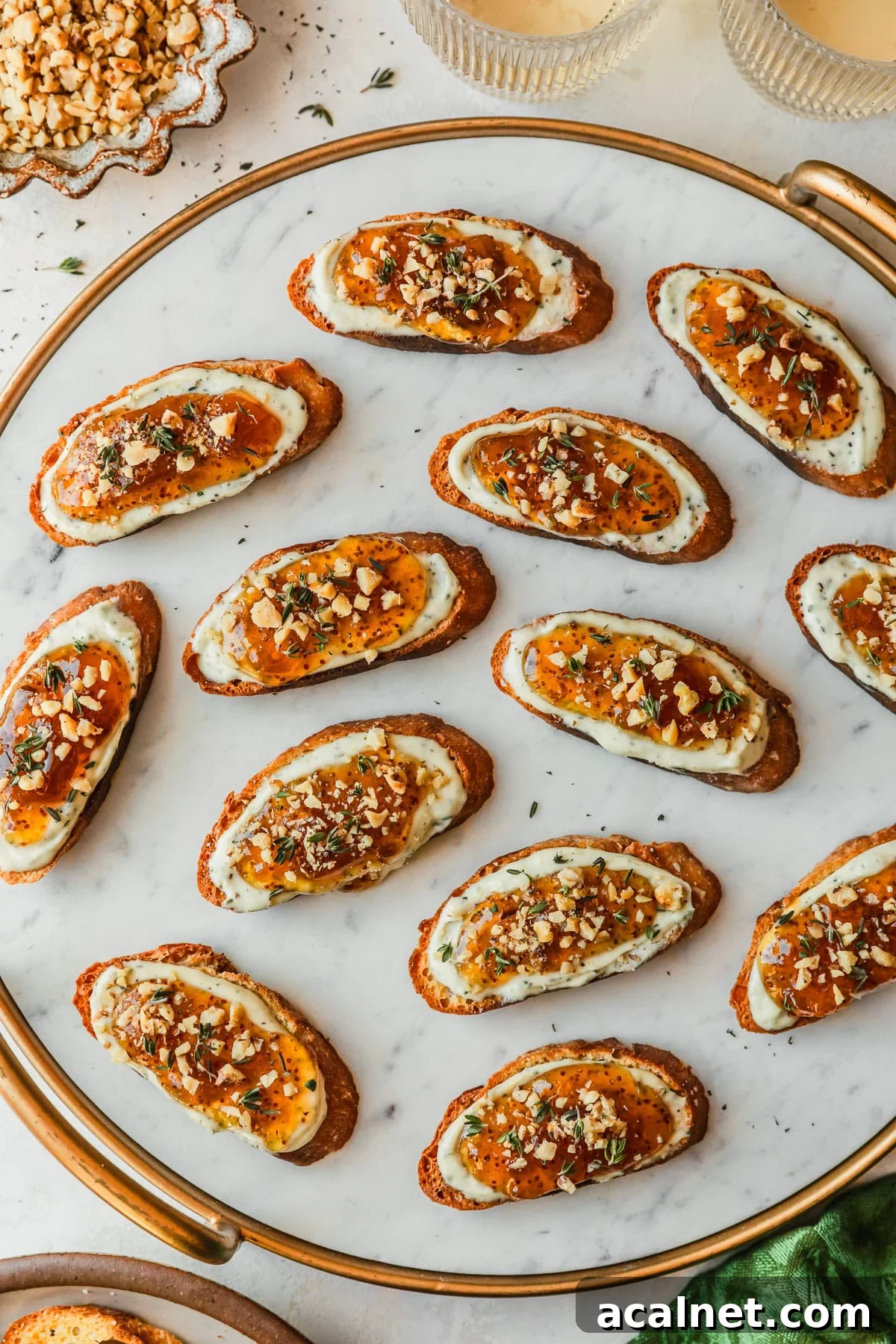 A marble platter with fig, honey, and goat cheese crostini on a white counter next to glasses of white wine, a green linen, and a brown bowl of walnuts.