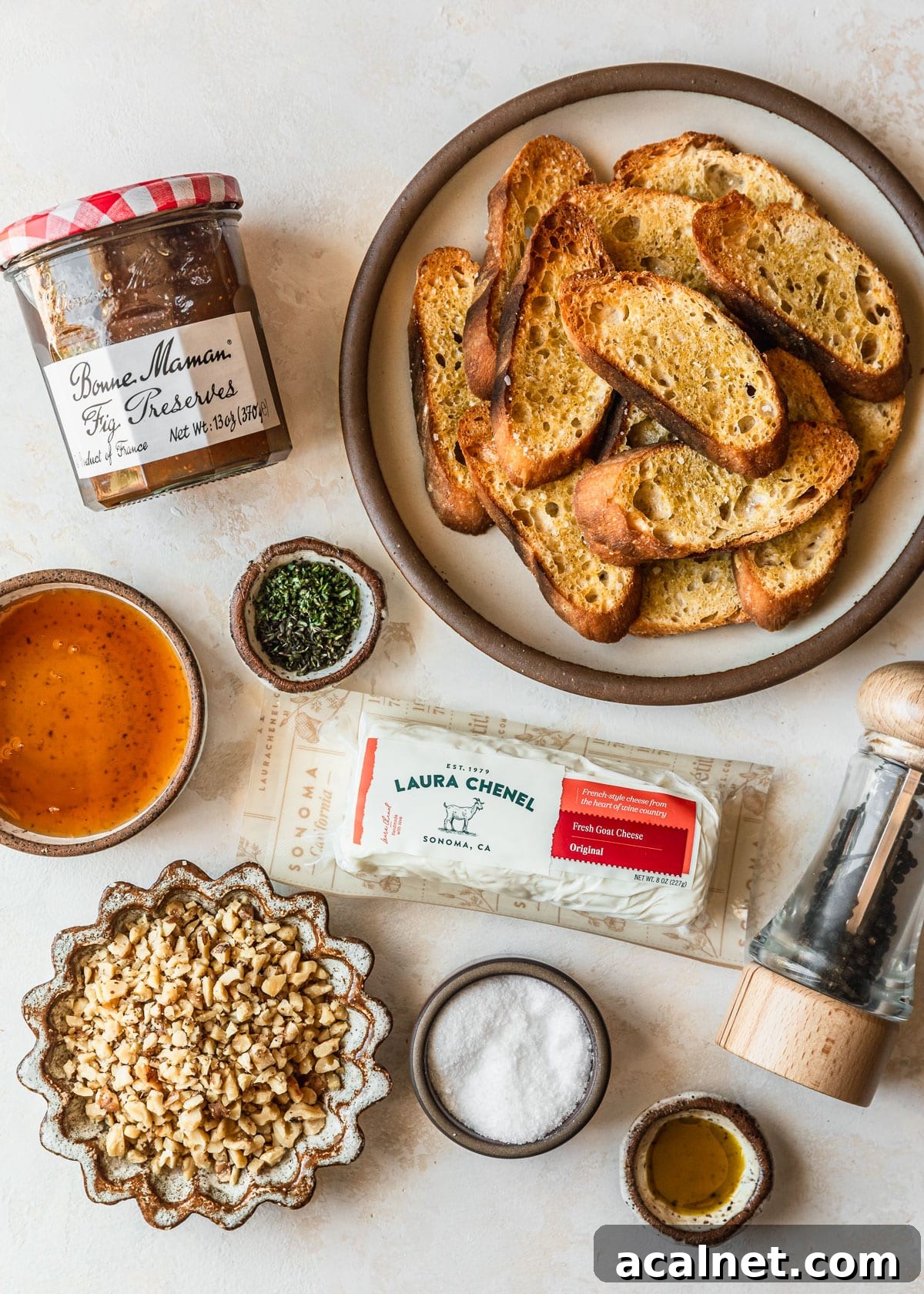 White and brown bowls of bread, jam, honey, herbs, salt, oil, walnuts, and cheese on a tan counter.