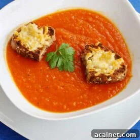 Creamy Roasted Butternut Squash and Tomato Soup Recipe 12 Closeup of a white dish of butternut squash and tomato soup with little cheese toasts on top on a bright blue background with a decorative spoon in front.