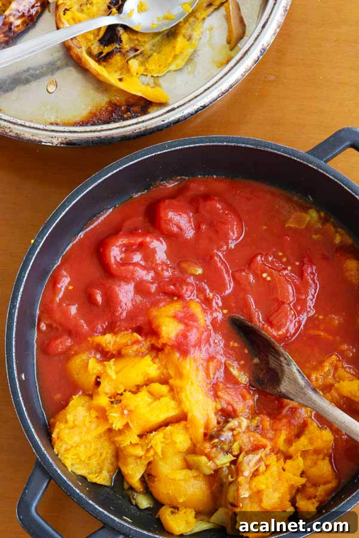 Creamy Roasted Butternut Squash and Tomato Soup Recipe 6 Mashed up roasted butternut squash and canned tomatoes in a black pan with a wooden spoon, with the empty roasted butternut squash skin visible at the side.