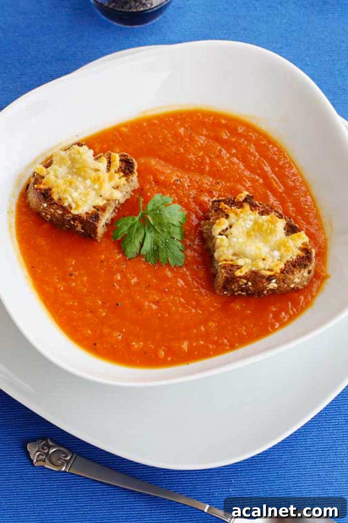 Creamy Roasted Butternut Squash and Tomato Soup Recipe 7 Closeup of a white dish of butternut squash and tomato soup with little cheese toasts on top on a bright blue background with a decorative spoon in front.