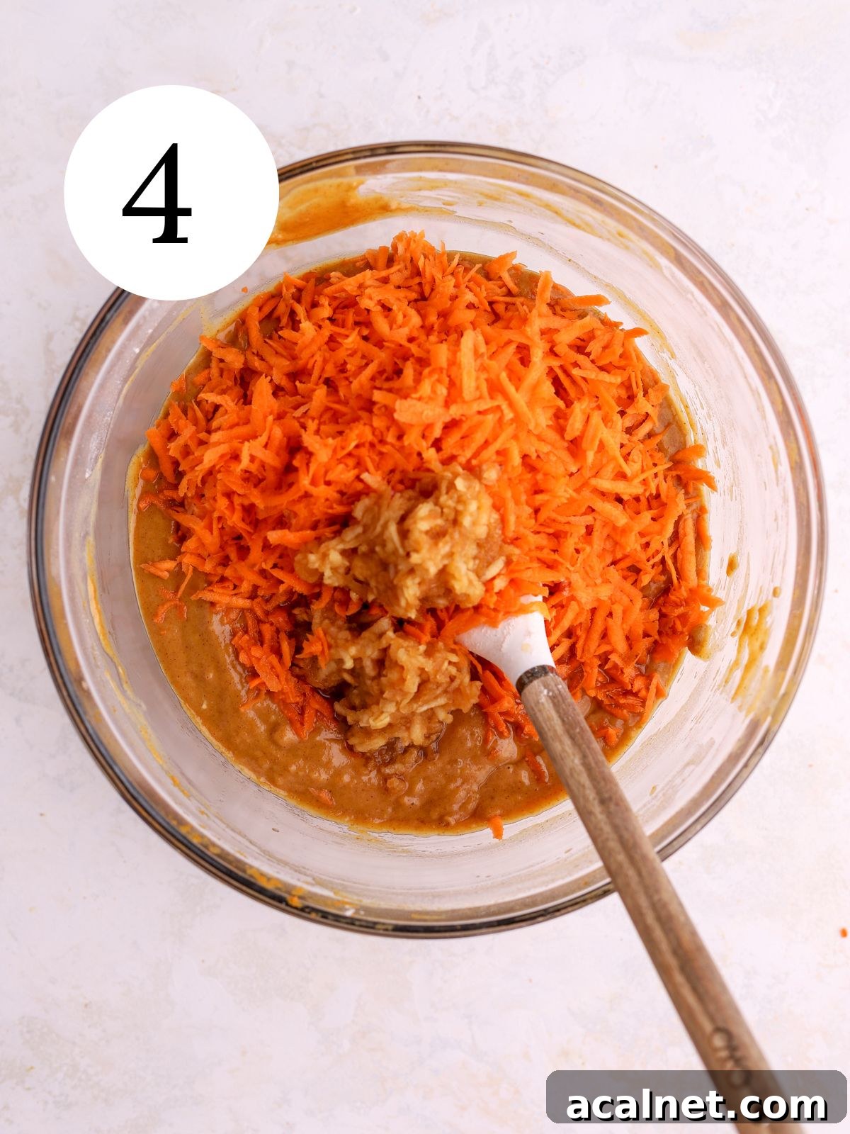 A mixing bowl with shredded carrots being stirred into pumpkin cake batter with a spatula. 