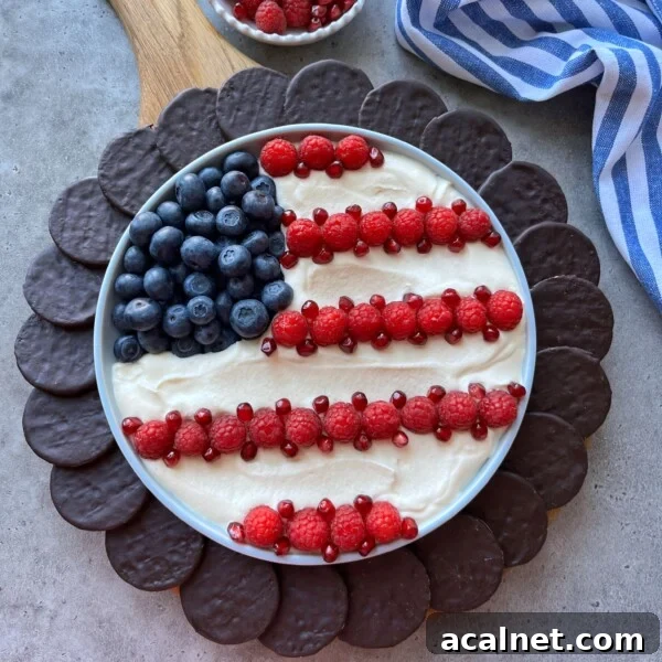 A round 4th of July dessert dip platter decorated like the American flag with blueberries, raspberries, and pomegranate seeds on a creamy base, surrounded by chocolate cookies. A striped blue and white cloth is nearby.