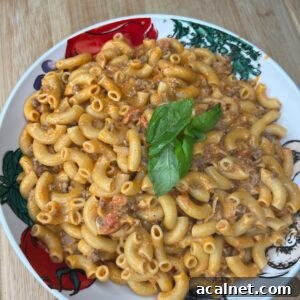 A plate of macaroni pasta with a creamy, tomato-based meat sauce, garnished with fresh basil leaves, served on a decorative plate with vegetable illustrations, on a wooden surface.