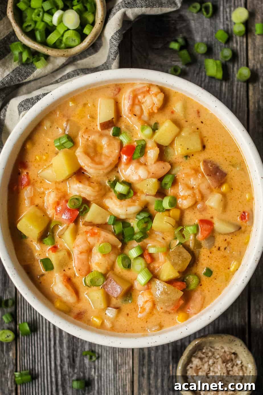 Creamy Cajun Shrimp and Sweet Corn Chowder Recipe 2 overhead shot of creamy cajun shrimp and corn chowder in a white bowl on a wooden surface. Bowl of salt and chopped green onions in top left and bottom right of shot.