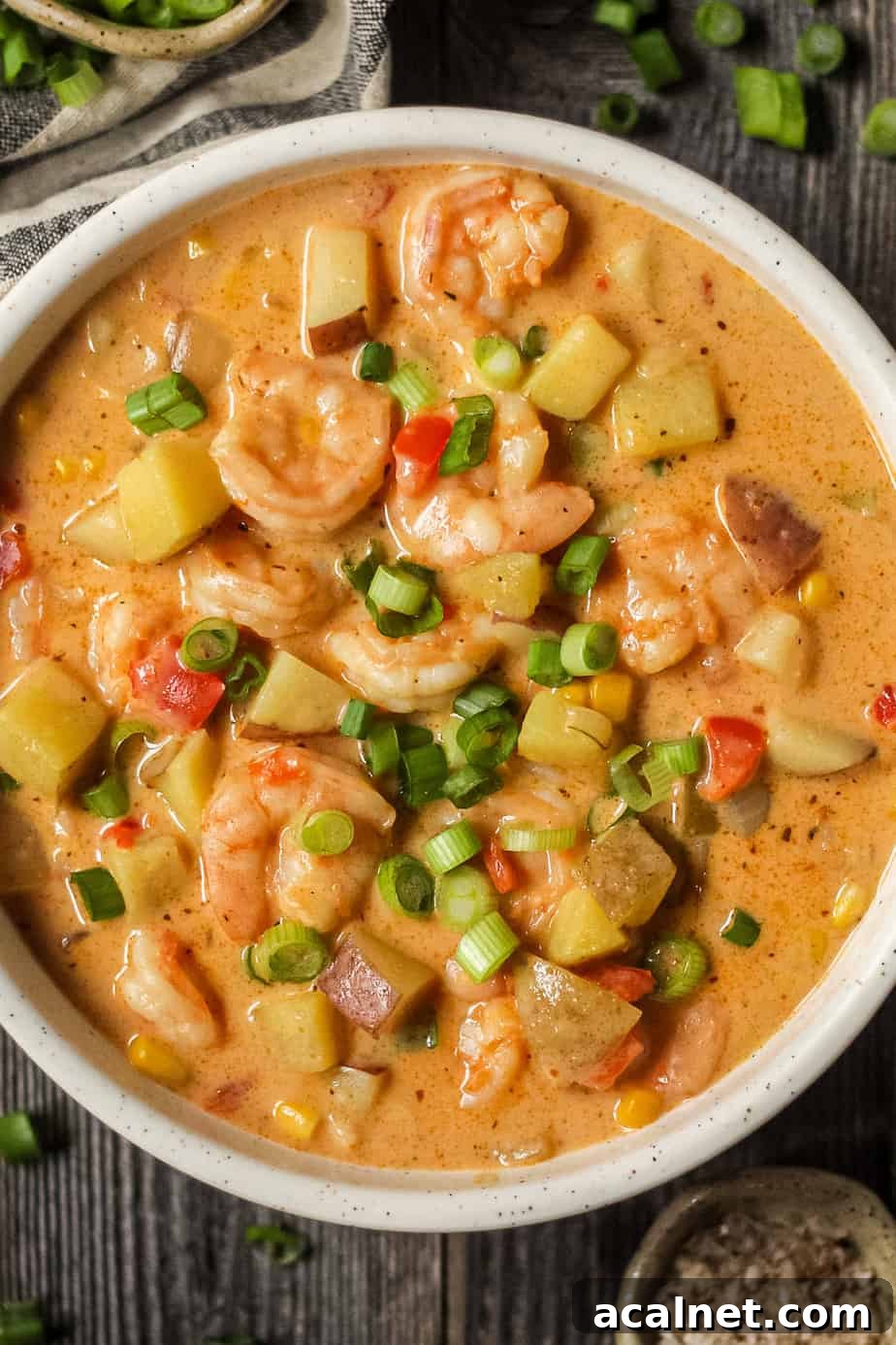 Creamy Cajun Shrimp and Sweet Corn Chowder Recipe 12 overhead shot of creamy cajun shrimp and corn chowder in a white bowl on a wooden surface. Bowl of salt and chopped green onions in top left and bottom right of shot.