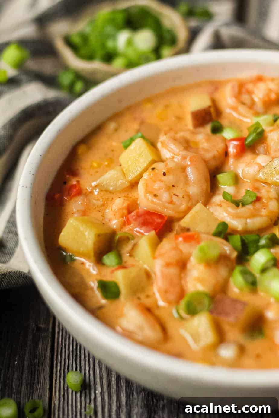 Creamy Cajun Shrimp and Sweet Corn Chowder Recipe 3 side angle shot of creamy cajun shrimp and corn chowder in a white bowl on a wooden surface. Bowl of salt and chopped green onions in top left and bottom right of shot.