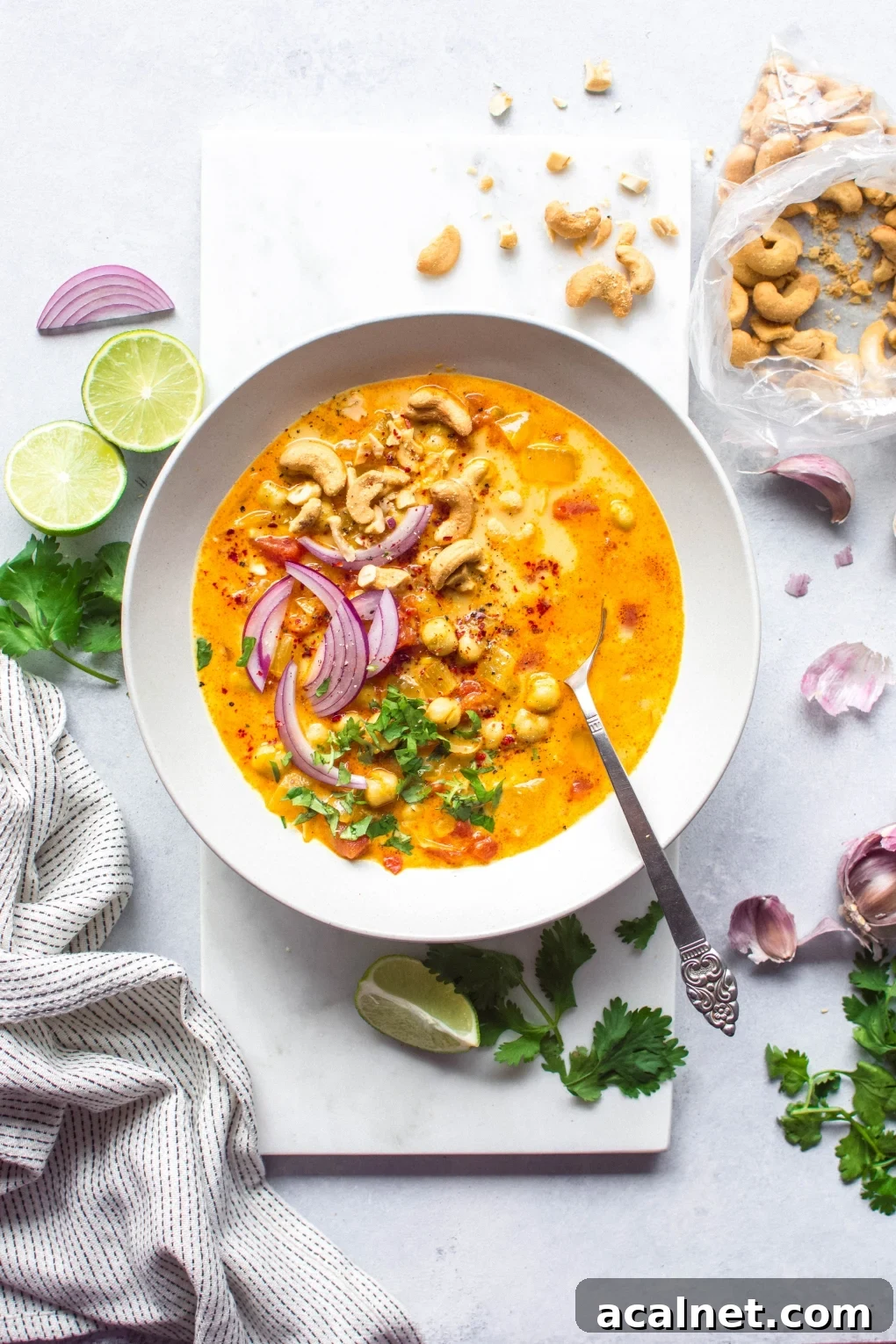 Large white bowl of bright orange chickpea and tomato coconut curry soup. Soup is topped with fresh red onion slices, chopped herbs, curry spiced cashews, and lime wedges. On a white background next to scattered herbs, lime wedges, and cashew bits.