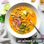Large white bowl of bright orange chickpea and tomato coconut curry soup. Soup is topped with fresh red onion slices, chopped herbs, curry spiced cashews, and lime wedges. On a white background next to scattered herbs, lime wedges, a soft striped dish towel, and cashew bits.