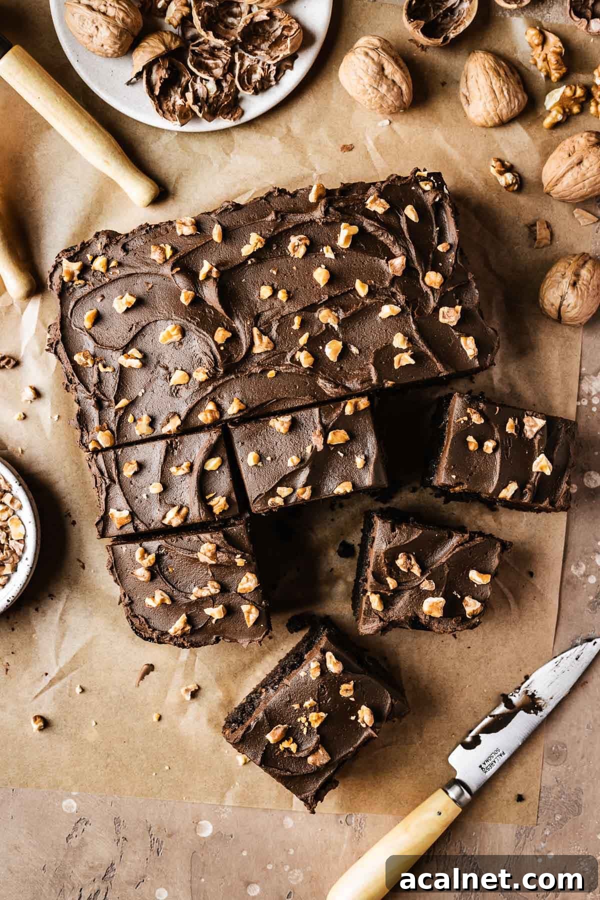 A single layer square chocolate cake topped with nuts on brown parchment paper.