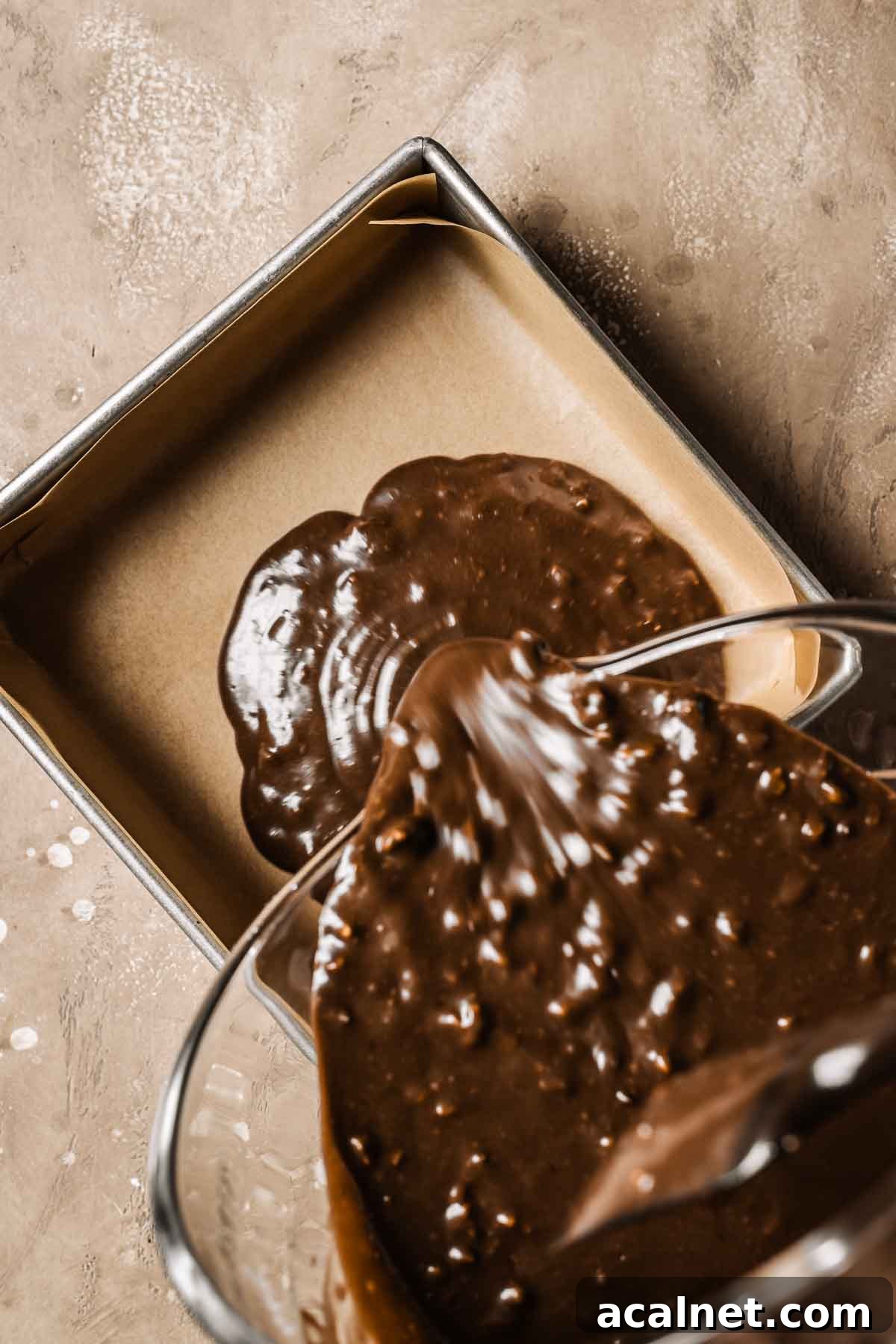 Chocolate walnut cake batter being poured from a mixing bowl into a parchment lined square pan.