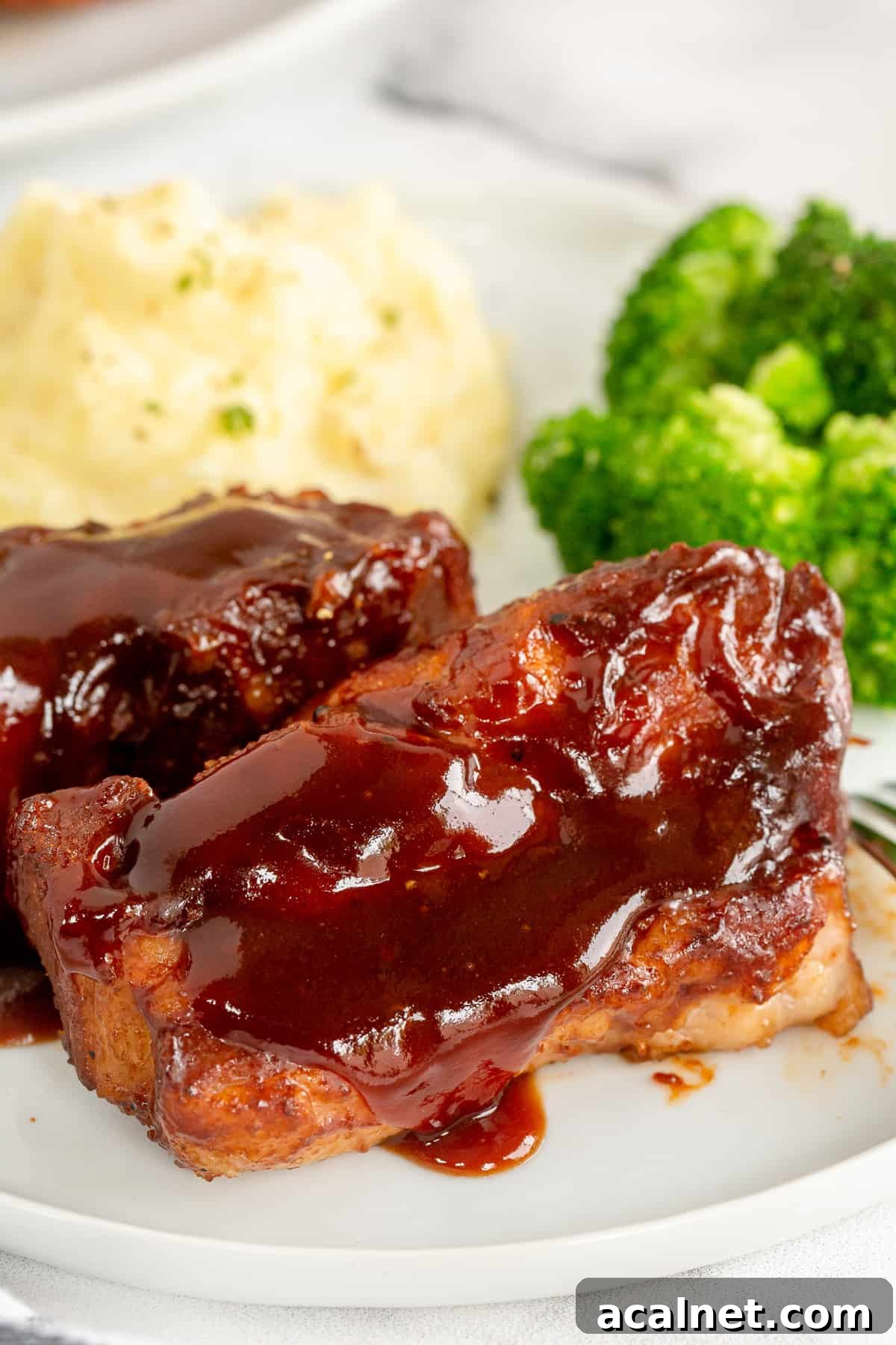 Two country style ribs with bbq sauce on a plate with mashed potatoes and broccoli.