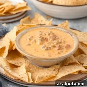 A bowl of creamy cheese dip with sausage and diced tomatoes in a small bowl on a large plate, accompanied by a plate of tortilla chips.