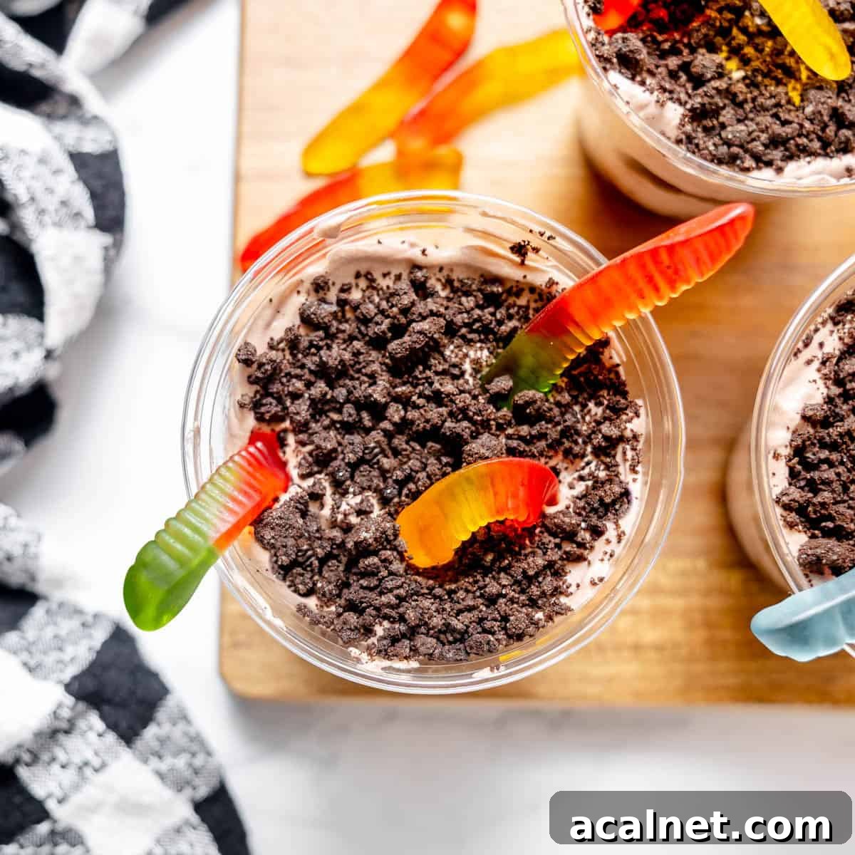 A cup filled with chocolate pudding, crumbled cookies, and gummy worms on top, placed on a wooden board next to a black and white checkered cloth.