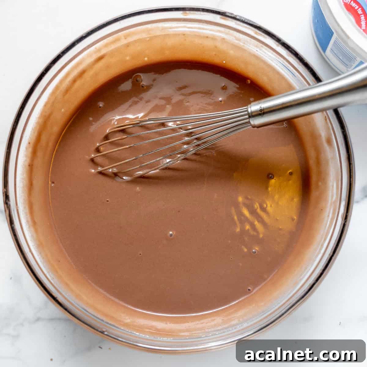A whisk resting in a glass bowl filled with smooth chocolate pudding on a white surface.