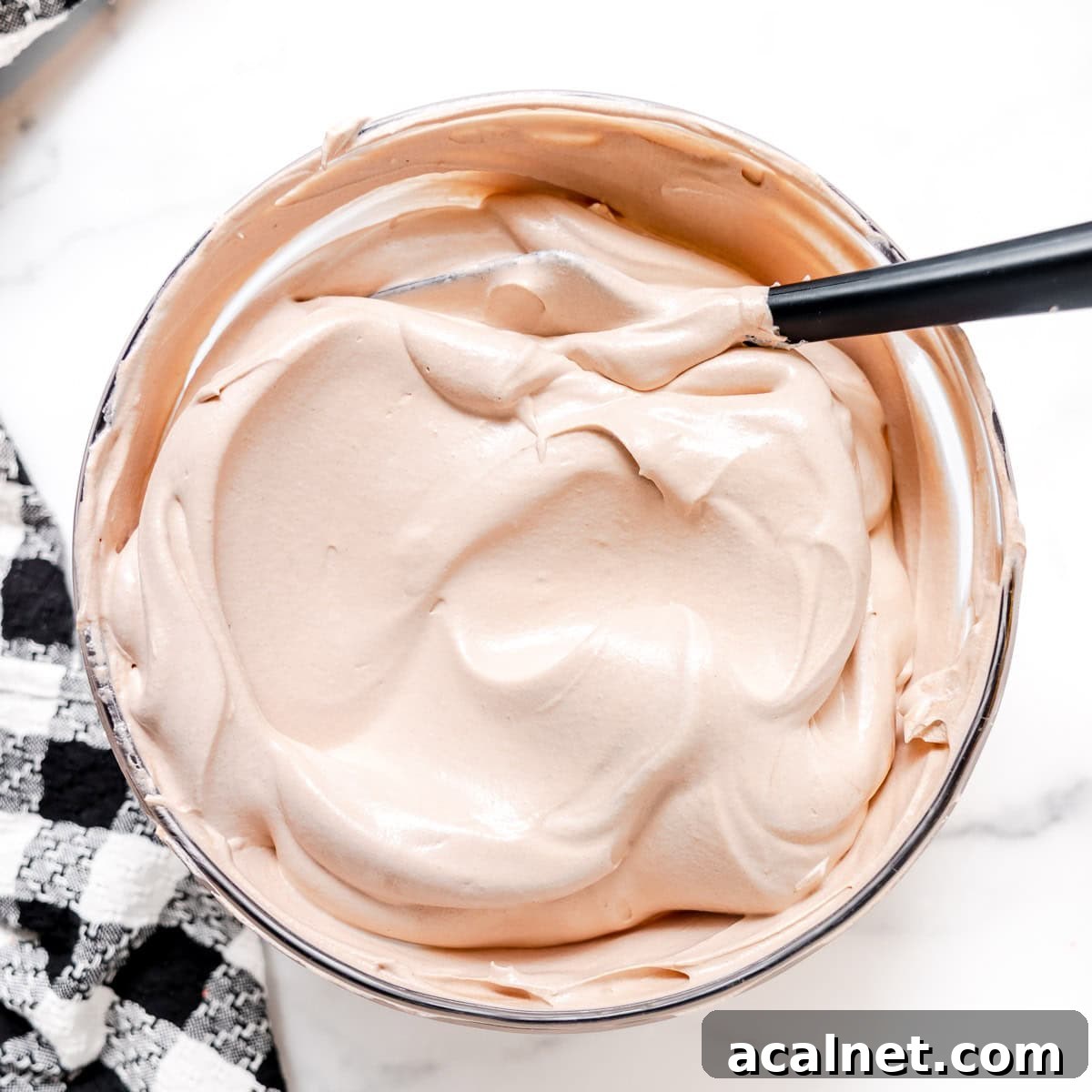 A bowl of light brown whipped mixture with a spoon resting inside, placed on a white surface next to a black and white checkered cloth.