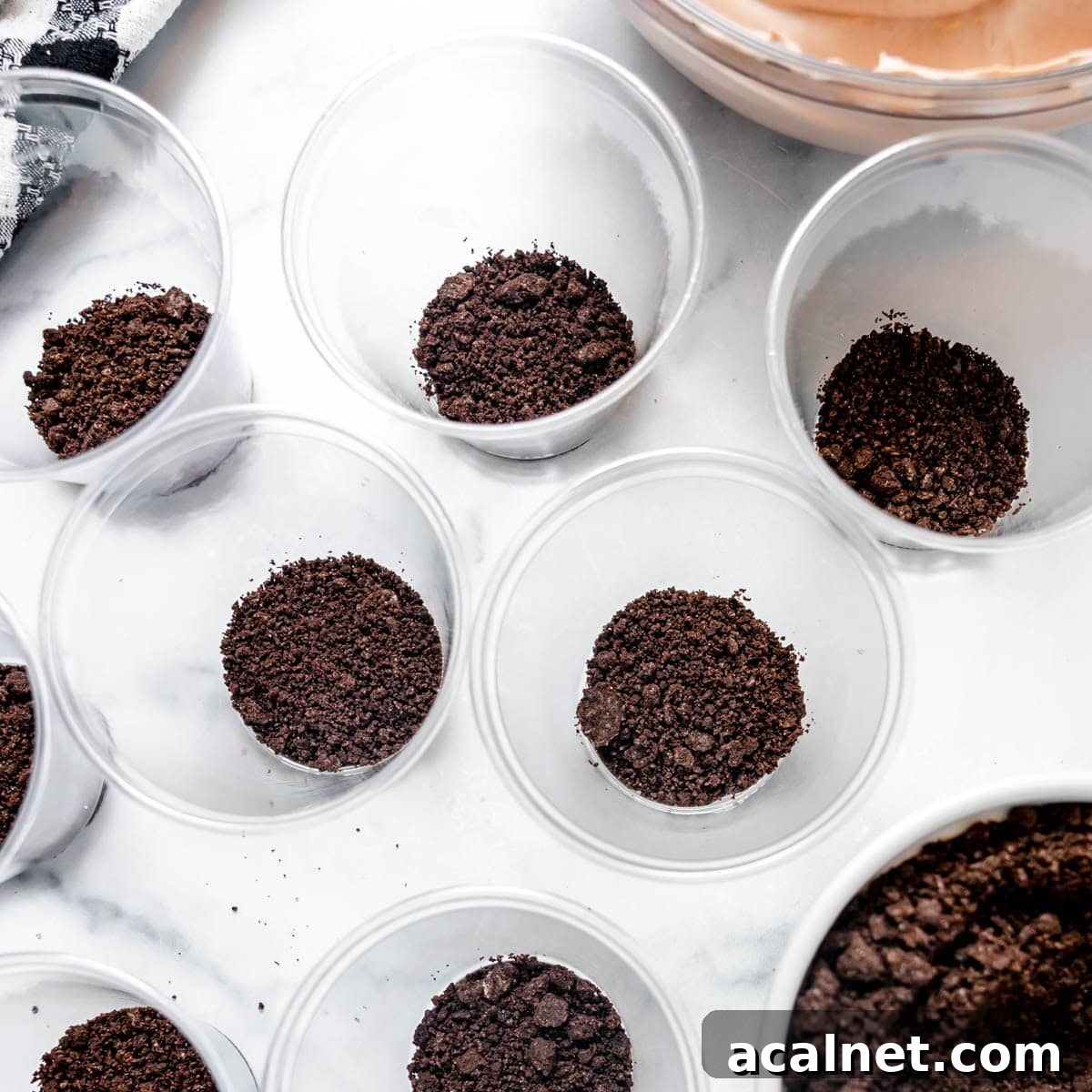 Several clear plastic cups are partially filled with a layer of crushed chocolate cookies on a white surface. A bowl with a brown mixture is partially visible in the corner.