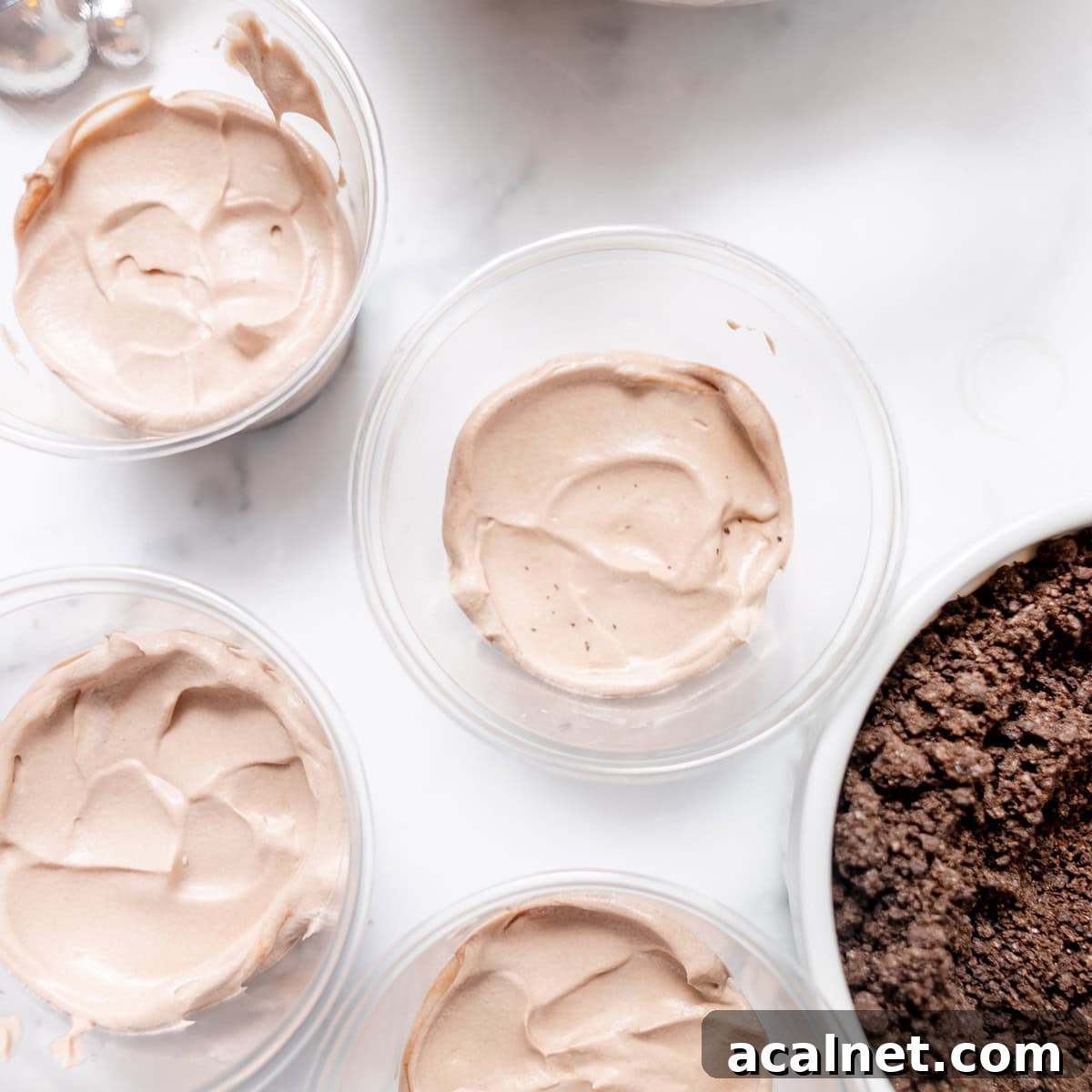 Four clear cups filled with light brown mousse sit on a marble surface next to a bowl of crumbled chocolate cookies.