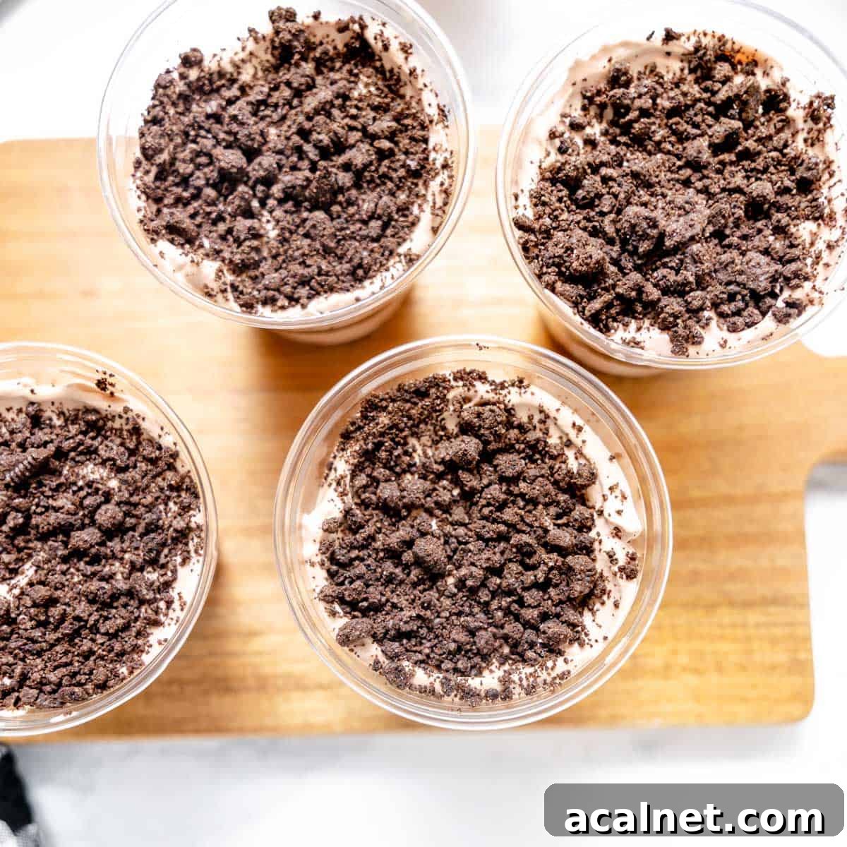 Four clear cups filled with chocolate mousse topped with crumbled chocolate cookies are arranged on a wooden board.